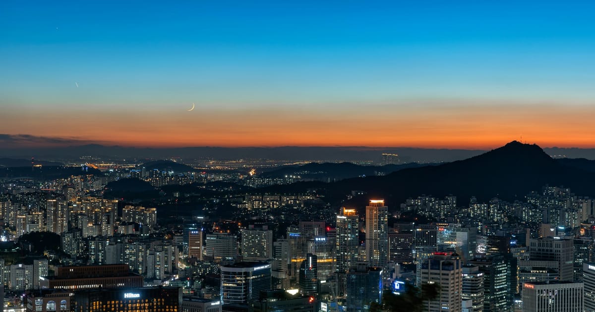 La silueta iluminada de Seúl al anochecer, con una luna creciente sobre las montañas.
