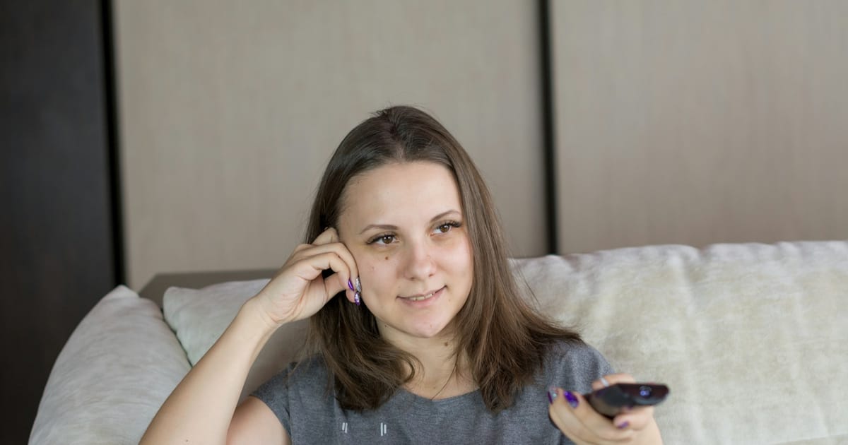 Una mujer sentada en un sofá sostiene un control remoto y sonríe.